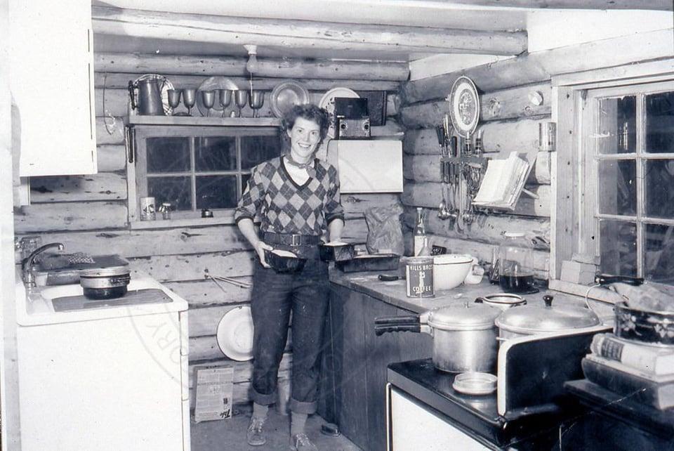 A woman with short curly hair, wearing jeans and an argyle sweater, holds two bread pans full of dough. She's in a small log cabin kitchen.