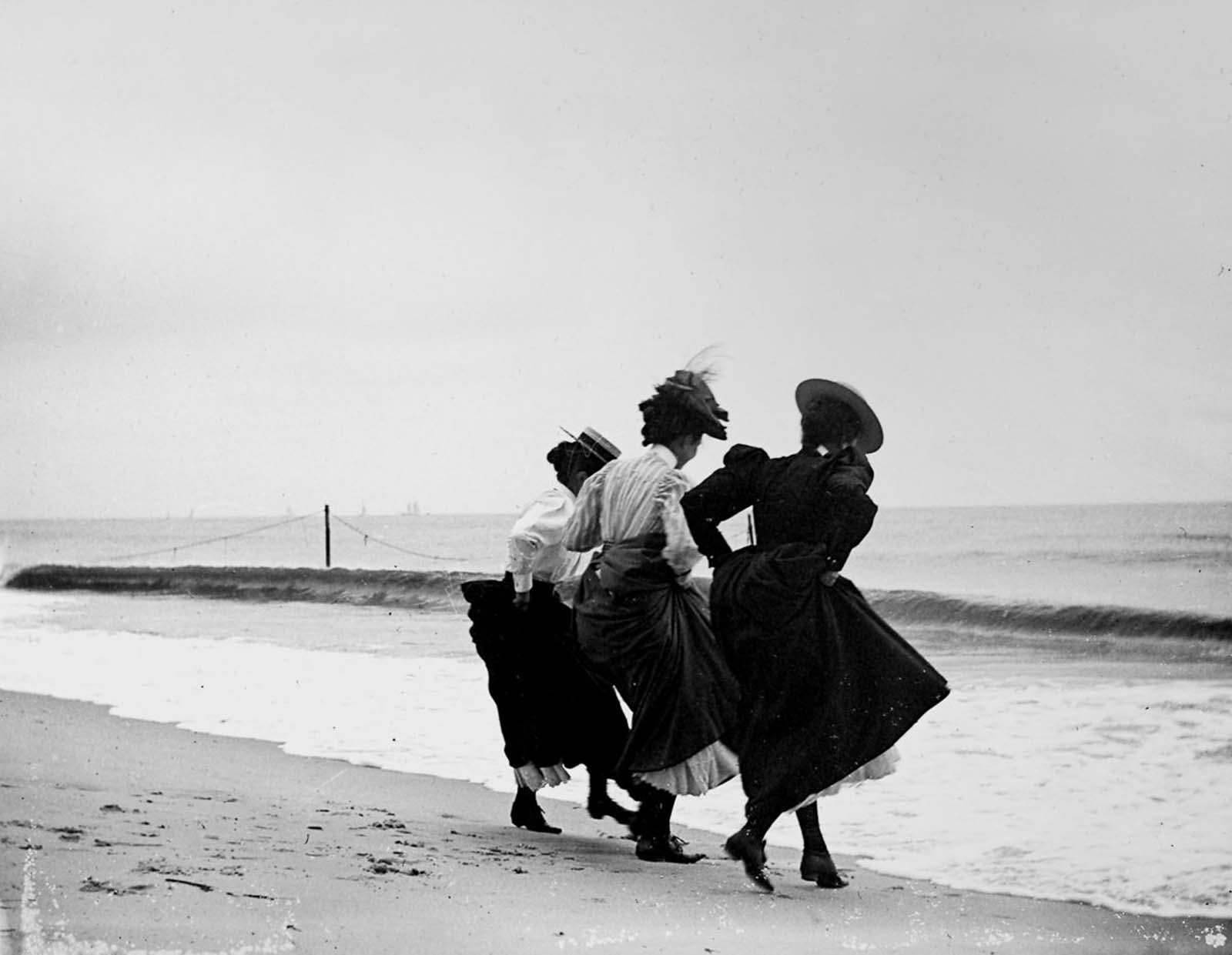 three Edwardian women hold their skirts up as they step towards the water on a beach