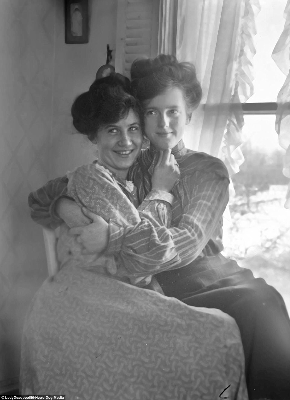 Two women sit in front of a window. The one on the right has her arms around the left one. The left one is touching the right's chin with her hand.