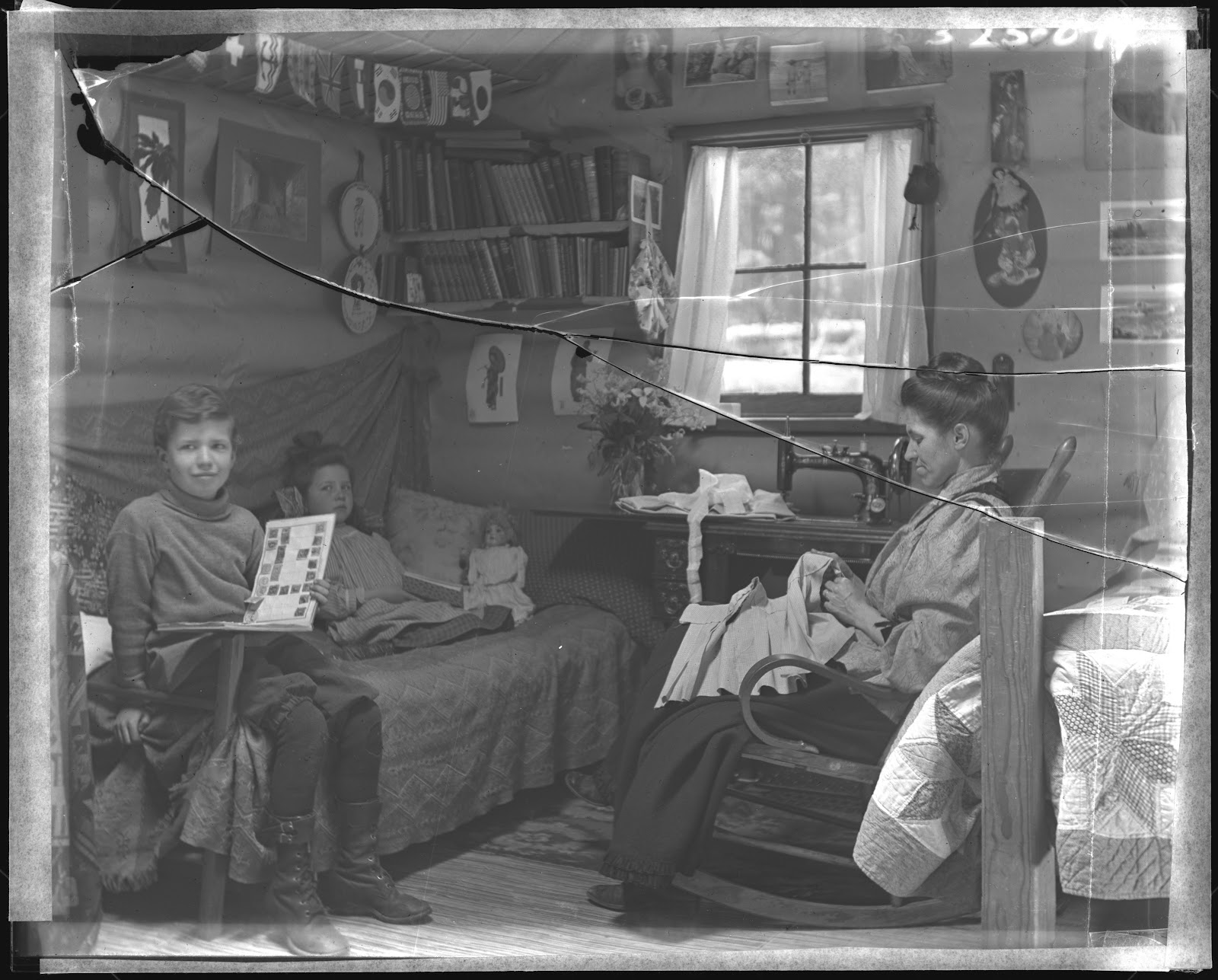 a woman sews in a rocking chair. Across from her are a young boy and girl sitting in a bed. The boy holds a book with squares in it, possibly photos or drawings. A doll is next to the girl, and there are shelves of books above the bed and framed wall decor.