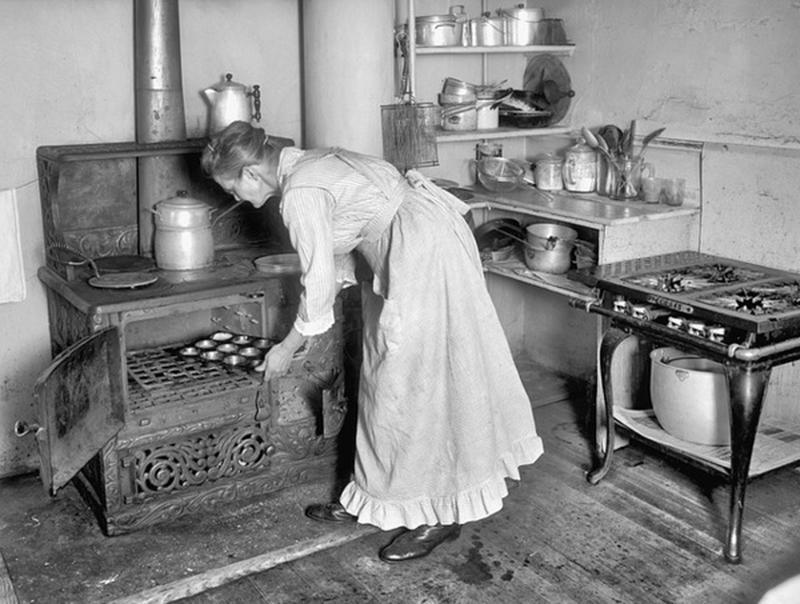 a woman in a long dreess bends over to put a muffin tray into a cast iron oven. Behind her is what looks like a gas range and a counter with pots and cooking utensils