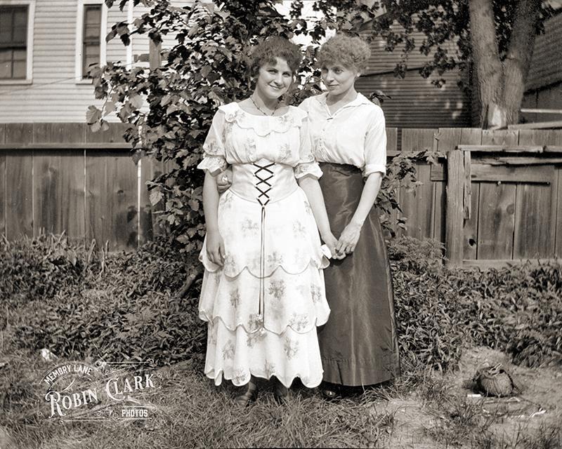 Two women with curly hair stand in a backyard. One is wearing a white floral dress with an underbust corset, the other a blouse and long skirt. They're holding hands.