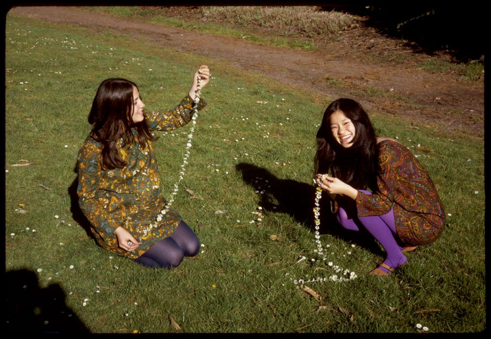 two young women, one white and one east Asian, kneel in the grass holding chains of white flowers. They're both wearing floral dresses in earthy colors and bright purple tights