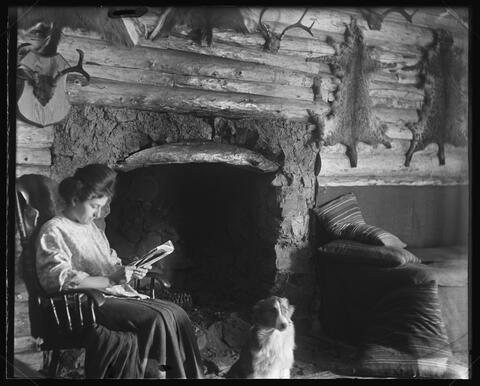 a young woman reads in a rocking chair in front of a fireplace in a log cabin, a dog sitting at her feet. The cabin's walls are decorated with furs and antlers.