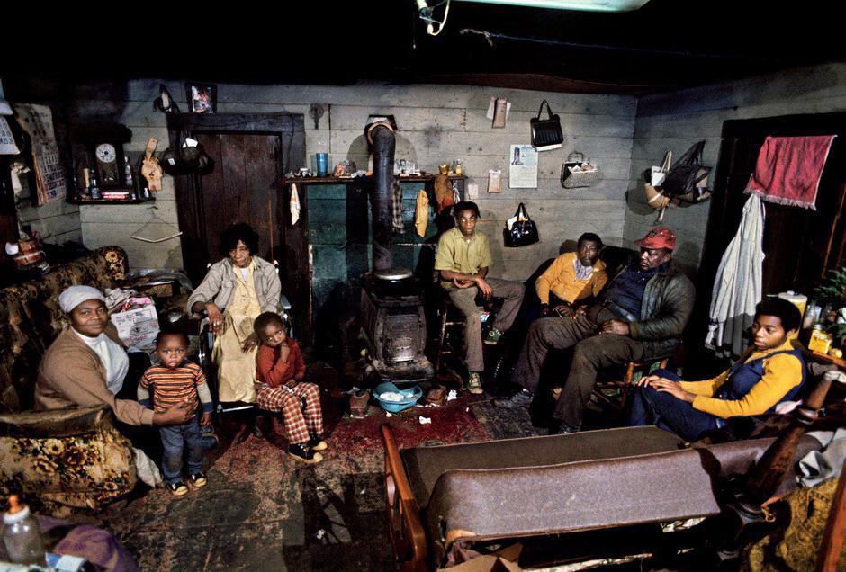 in a small room with wooden walls and a small cast iron stove sits a Black family on sofas and armchairs. From left to right is a woman with a hand on a small boy, an old woman with another small boy in front of her, two young men next to an older man with a red baseball cap, and another young man.