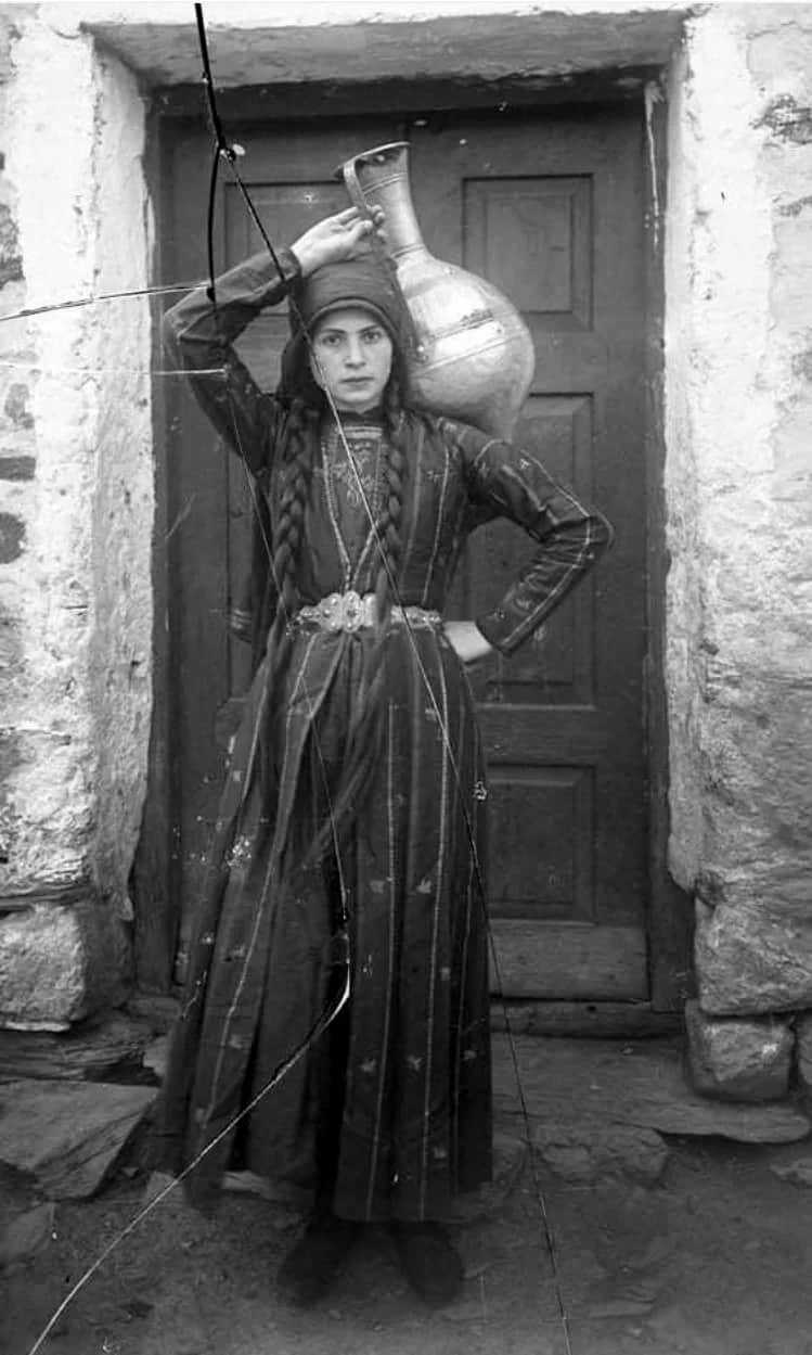 A young woman with long dark braids, a headscarf, and dark striped dress holds a large amphora propped on her shoulder.