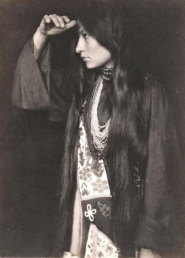 A young woman with long dark hair and many bead necklaces raises her hand to shade her eyes.