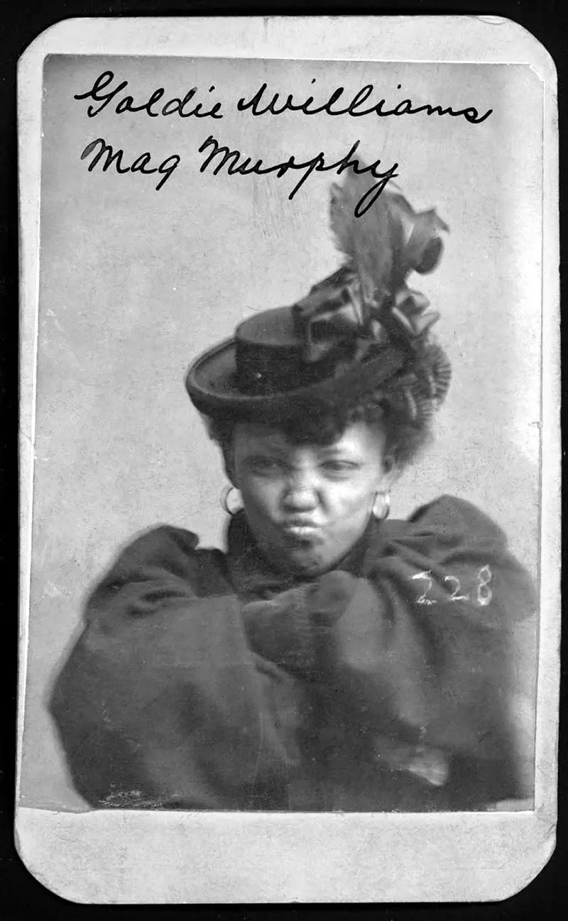 a young black woman wearing a hat decorated with ribbon and feathers makes an exaggerated pouting face, her arms crossed