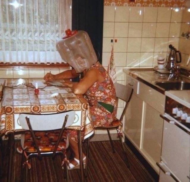 In a kitchen with tiled walls, a woman sits at a table writing on paper. Her head is covered with an inflated plastic hairdryer