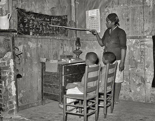 a Black woman uses a long stick to point at a wall hanging which has a cursive alphabet and the words 'The rain are fallin' written on it. Two Black children sit on chairs in front of it, watching.