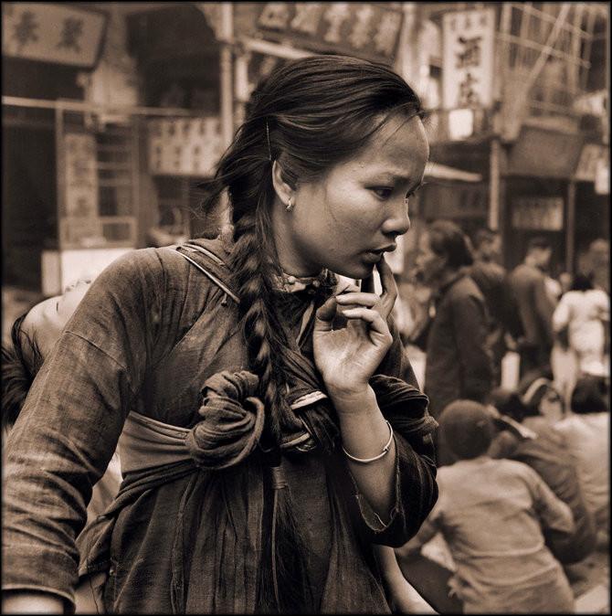 An east Asian woman looks like she's contemplating something, touching her finger to her mouth. Her baby's head is visible from behind her shoulder, and a sash is knotted in front of her chest.