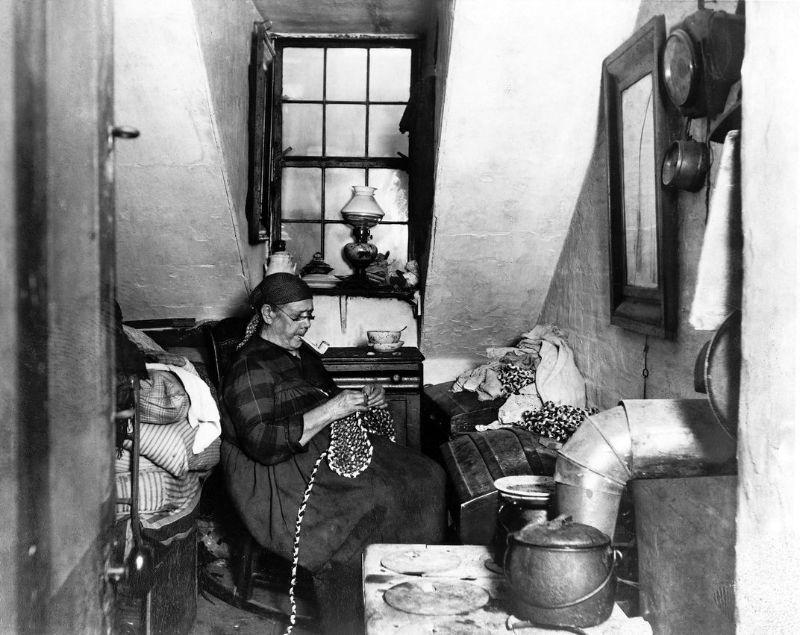 an old woman with glasses and a pipe sits in a tiny, cluttered room and is concentrating on a beaded object in her hands