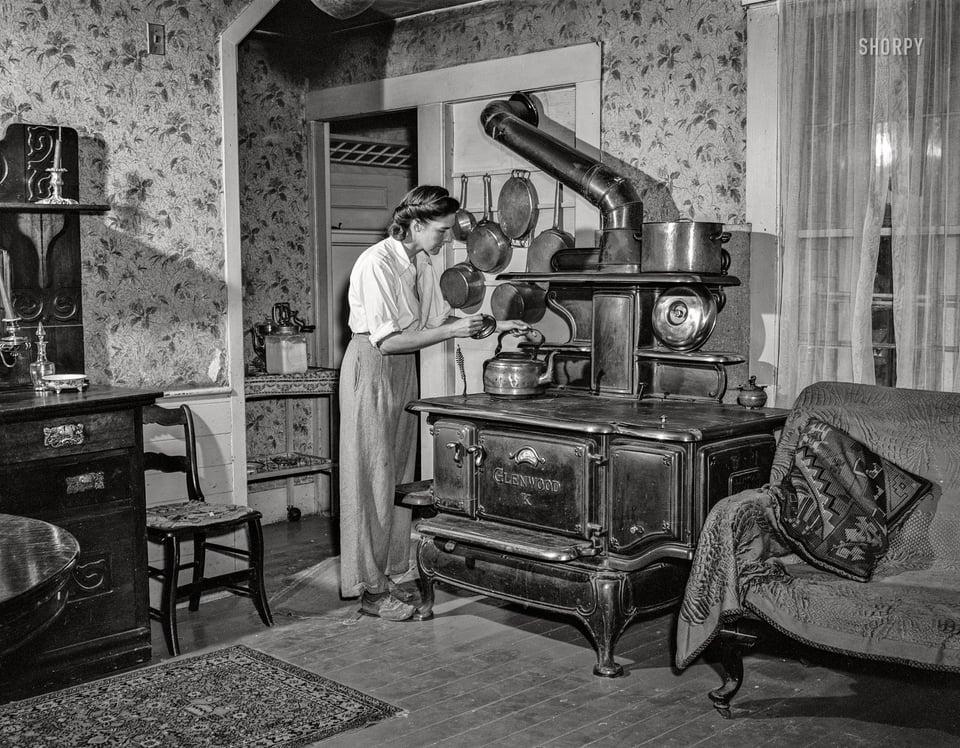 a woman in a home with floral wallpaper is placing a skettle on a cast iron stove.