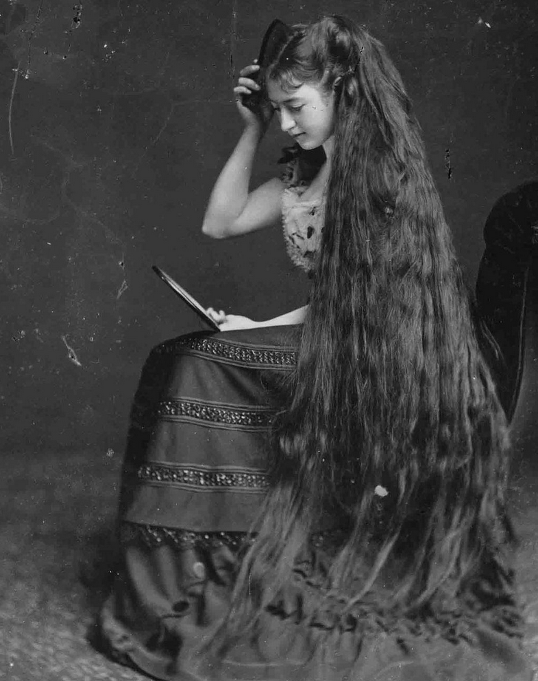 a seated young woman has long, thick dark hair that reaches to the floor. She holds a mirror as she combs her hair.