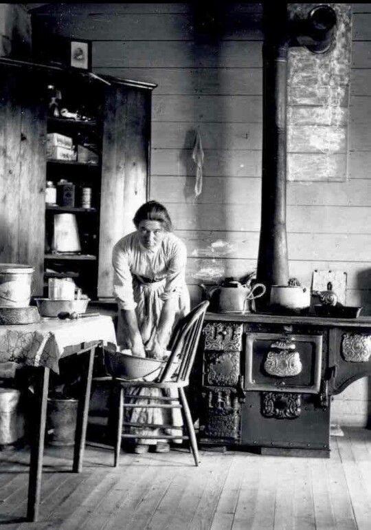 a woman faces the camera, bent over a large bowl on a chair. A cast iron stove is behind her, and the floor and walls are made of bare wood planks