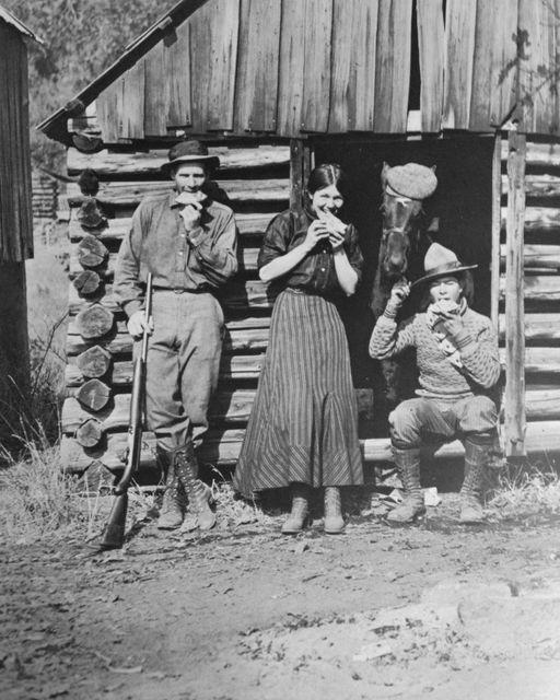 two young men, one holding a rifle, and a young woman pose in front of a log cabin while holding pieces of pie up to their mouths. Behind them, in the doorway, is a horse wearing a hat