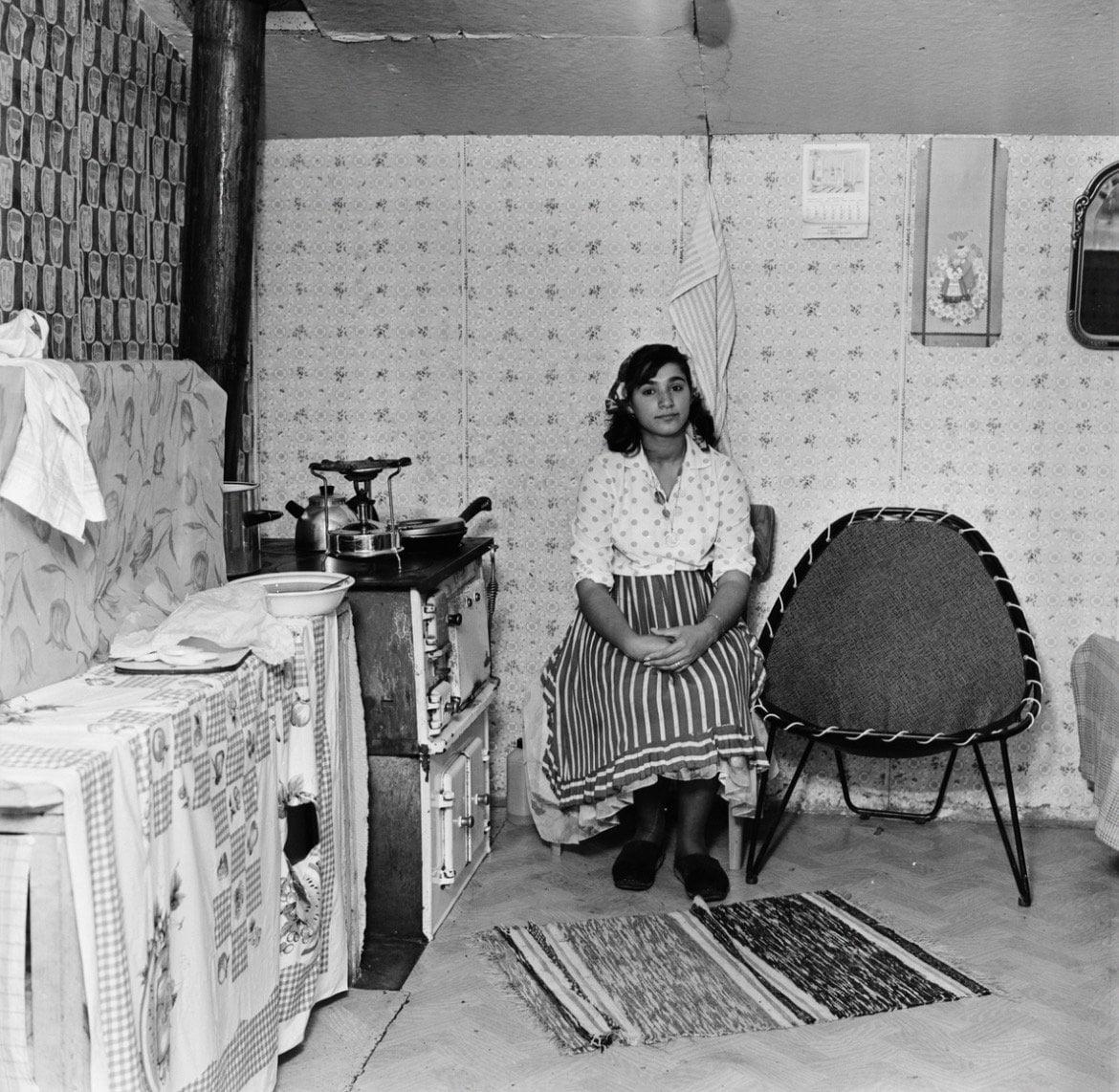 a young woman with a striped apron sits in front of a stove, facing the camera.