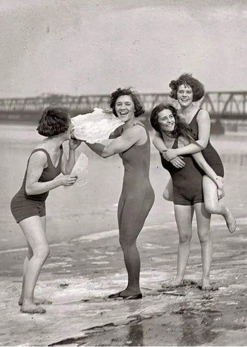  four women in bathing suits laugh while standing in shallow water on a beach. Two are holding what appears to be a sheet of ice, and one is riding piggyback on another