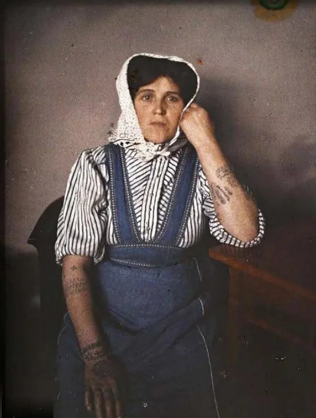 (in color). A woman wearing a headkerchief sits with her head propped by one hand, showing off small black tattoos on both her forearms.