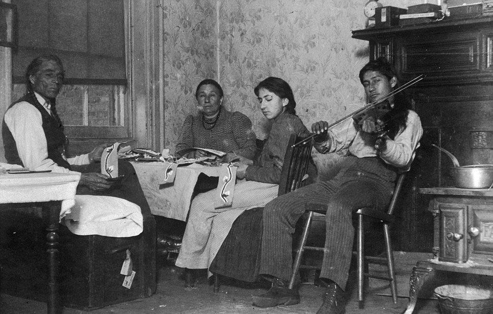 an older man and women with a younger man and woman sit around a table working with fabric in a cramped room. The young man is playing a violin or fiddle.