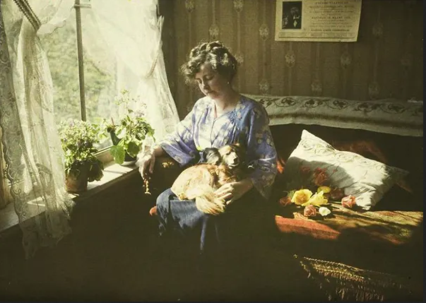 (in color). A woman sits on a couch in front of a window with lace curtains, the light softly shining through. She has a little fluffy dog in her lap.