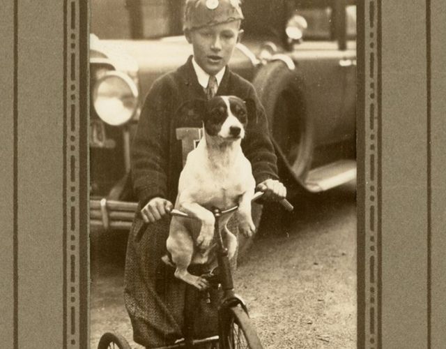 A young boy stands on a scooter, a dog sitting in front of him with its paws over the handlebars