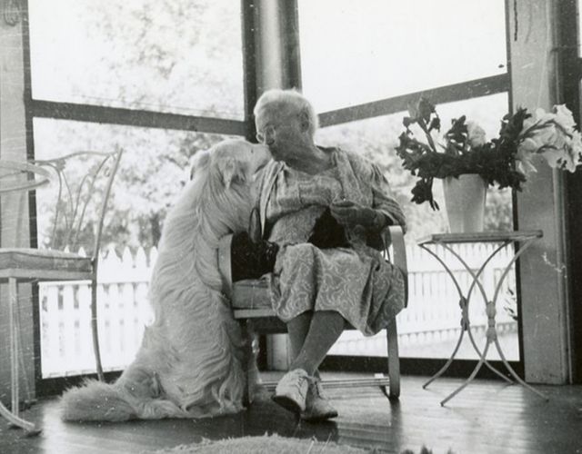 An old woman sits on a chair and kisses a large fluffy white dog on the nose