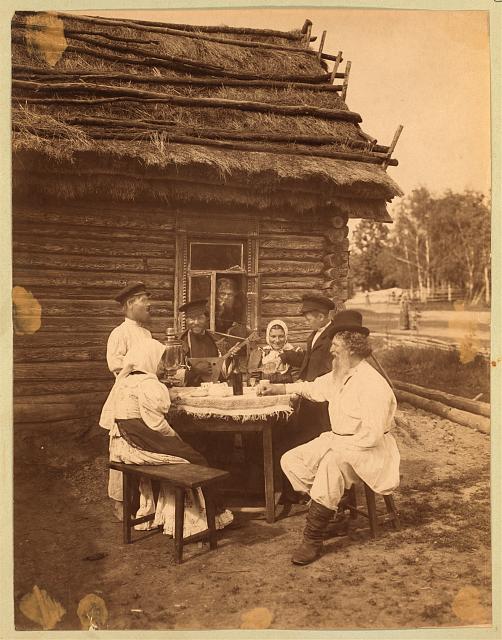 Five peasants sit around a table outside of a wooden house with a thatched roof.