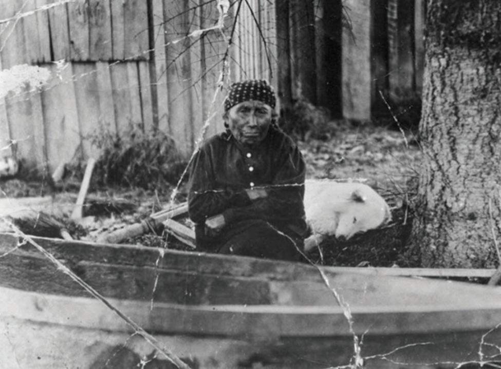 An old woman sits with a grim face and her arms crossed in front of a canoe. A medium-sized white dog sleeps behind her