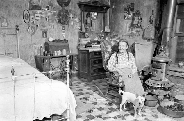 An older woman with two long braids sits in a rocking chair in front of a small woodstove. A little white dog stands in front of her. There's a large bed to the left, and behind her is a dresser with a mirror and a desk. The walls are covered in unframed pictures.