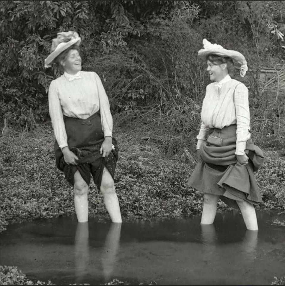 two women laugh at each other as they hold their skirts over their knees, standing in water that goes up to mid-calf